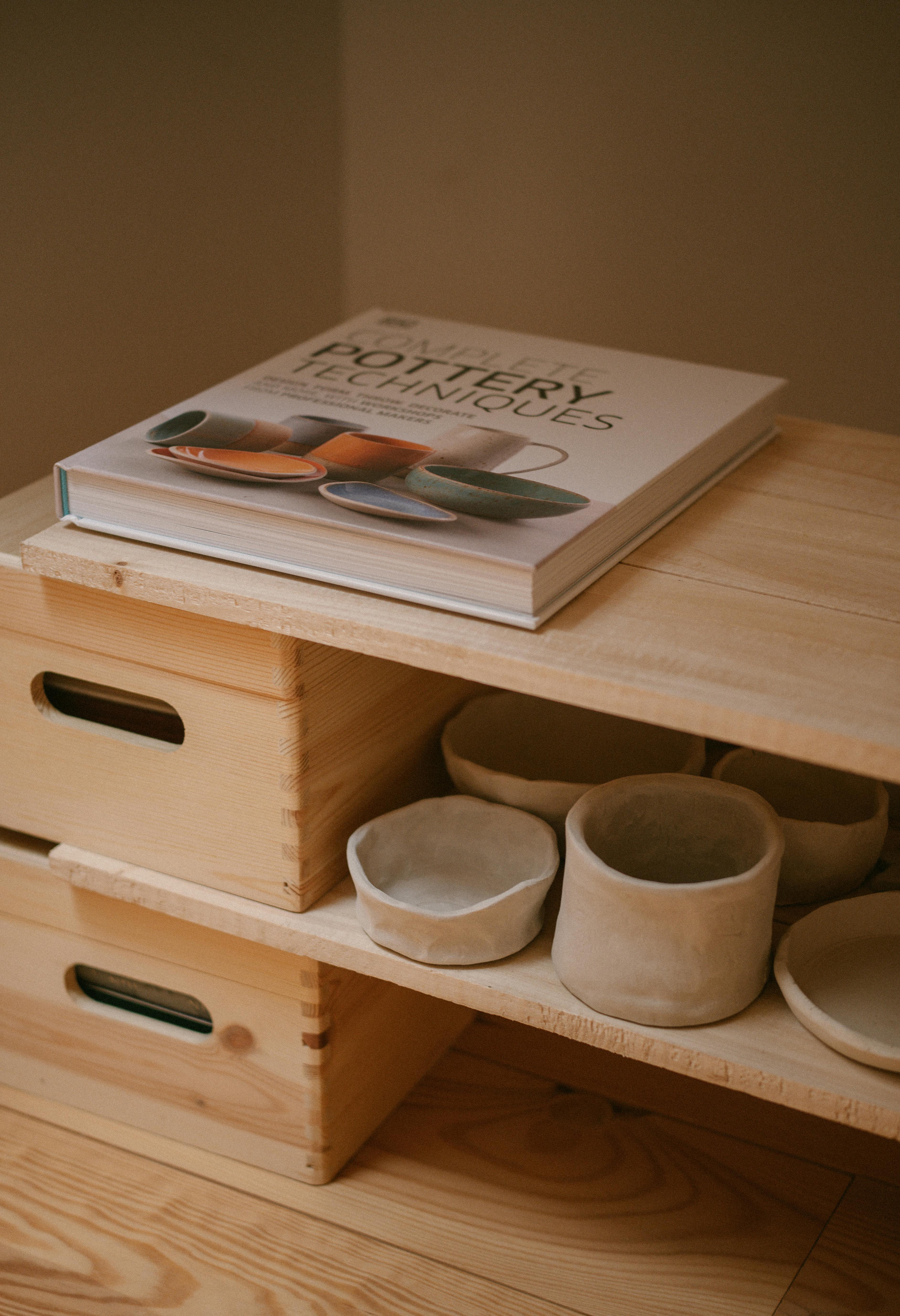 Aesthetic wooden shelves showcasing handmade pottery and a pottery techniques book in a cozy indoor setting.