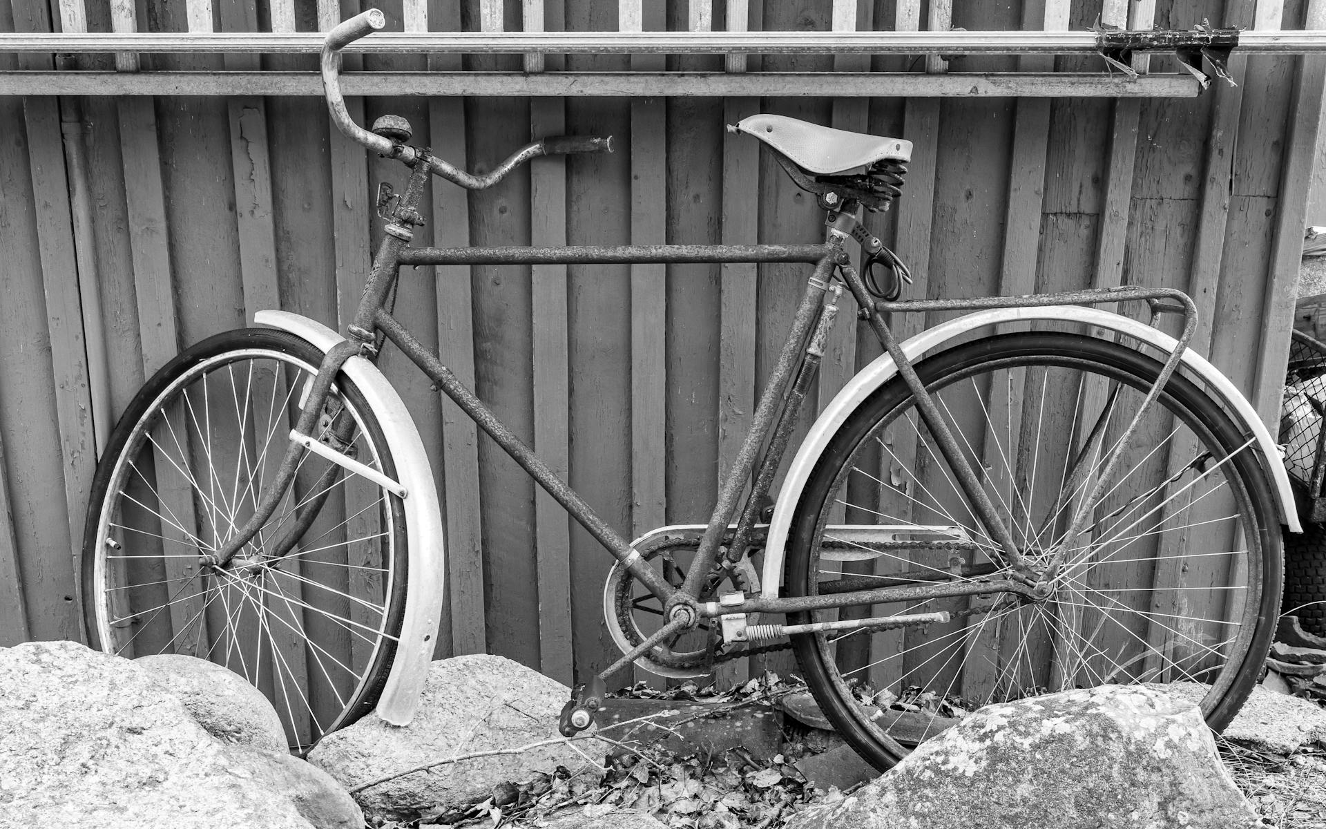 Monochrome photo of an old rusty bicycle leaning against a wooden fence outdoors.