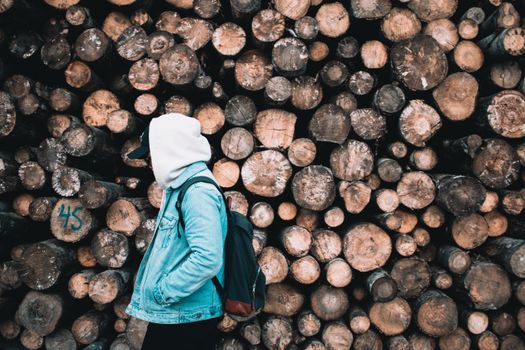 A person in a hoodie and jeans stands beside a large stack of wooden logs outdoors.