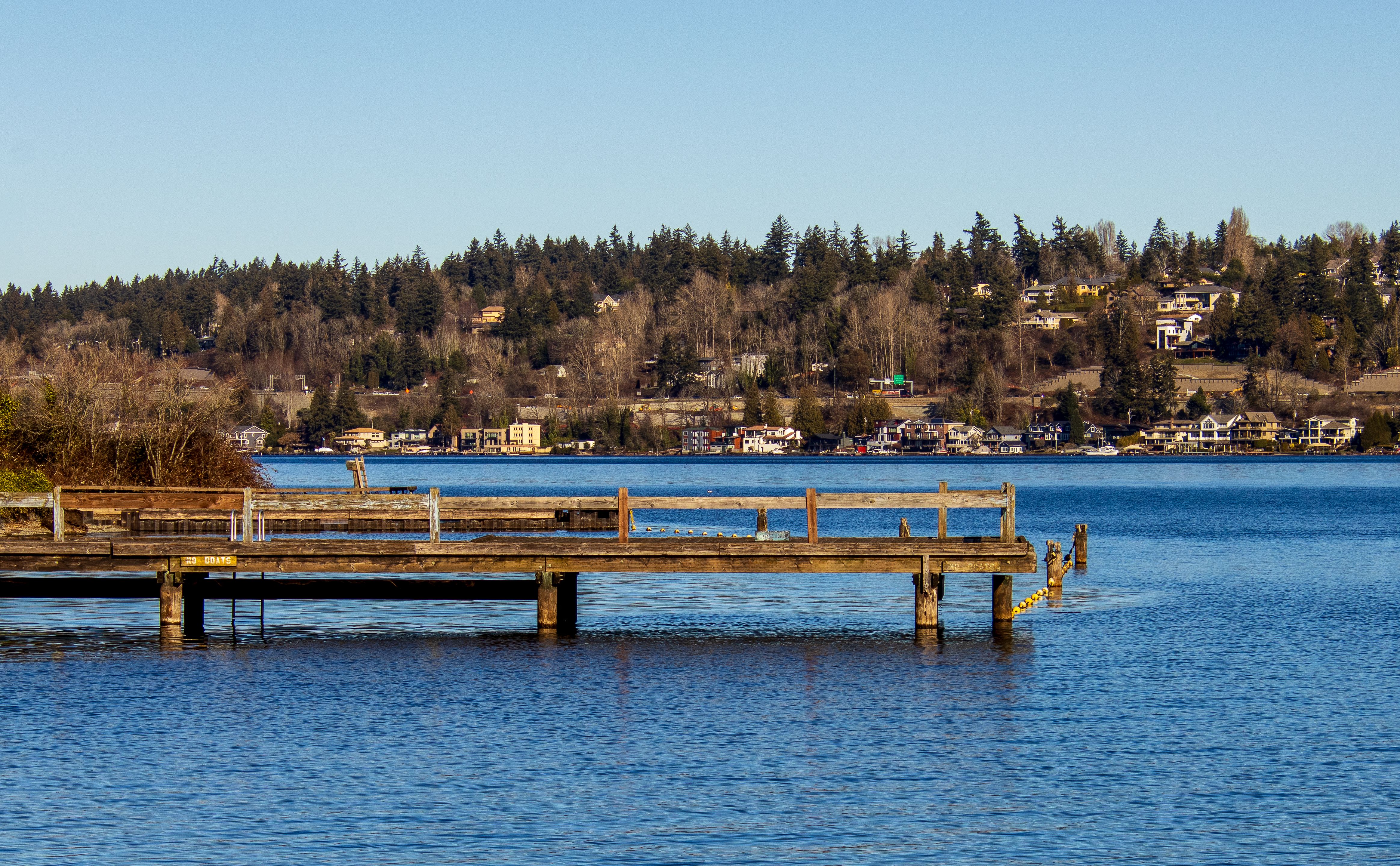 Scenic view of a calm lake with a wooden pier and houses in the background under a clear blue sky.