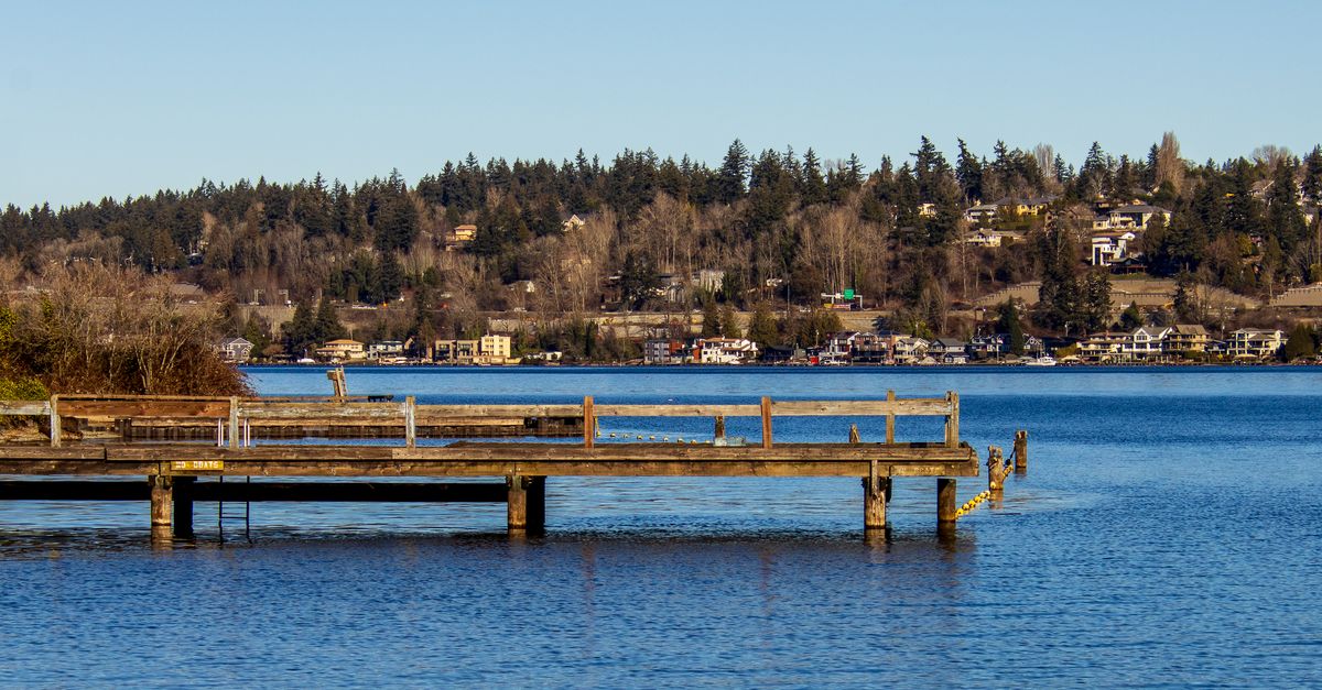 Scenic view of a calm lake with a wooden pier and houses in the background under a clear blue sky.