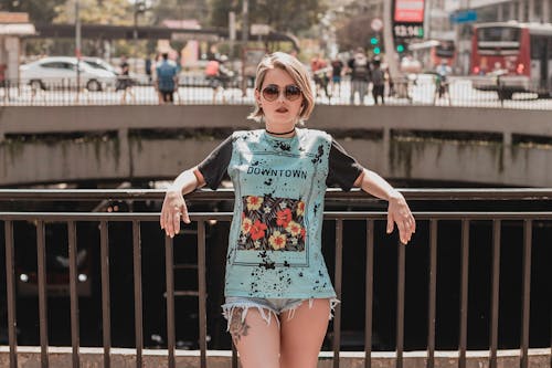 Ücretsiz Metal Korkuluk Pozuna Yaslanmış Güneş Gözlüğü, Tişört Ve Kot şortlu Kadın Fotoğrafı Stok Fotoğraflar