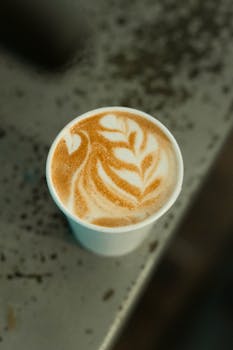 A beautifully crafted latte with intricate foam art served in a white paper cup on a textured surface.