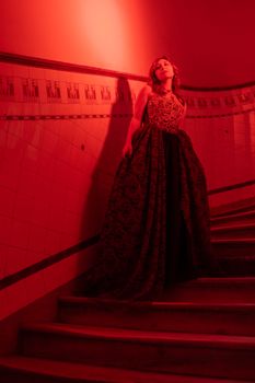 A woman poses in an ornate gown on a dimly lit staircase, bathed in red light.