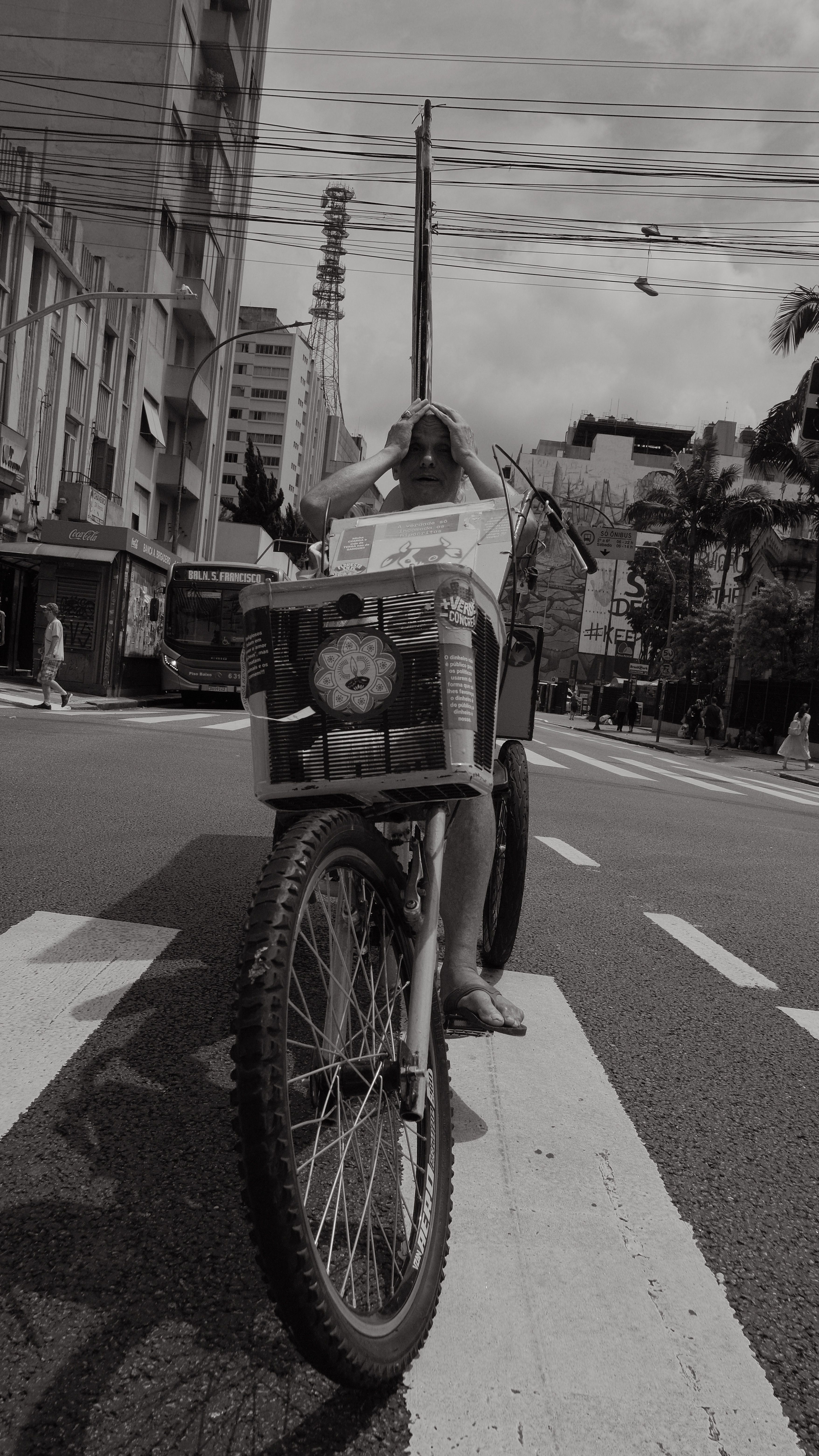 A black-and-white street photograph of a man reading a newspaper while sitting on a bicycle in a city.