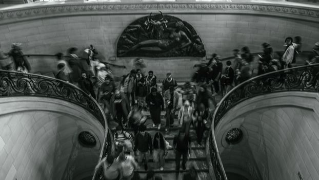 A bustling crowd descends a spiral staircase in Paris, captured in motion blur.