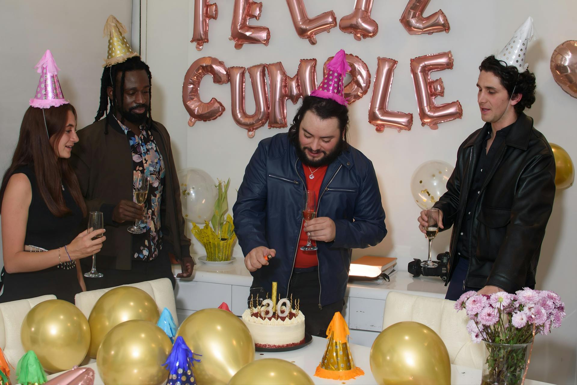 Group of friends celebrating a birthday with cake, balloons, and party hats indoors.