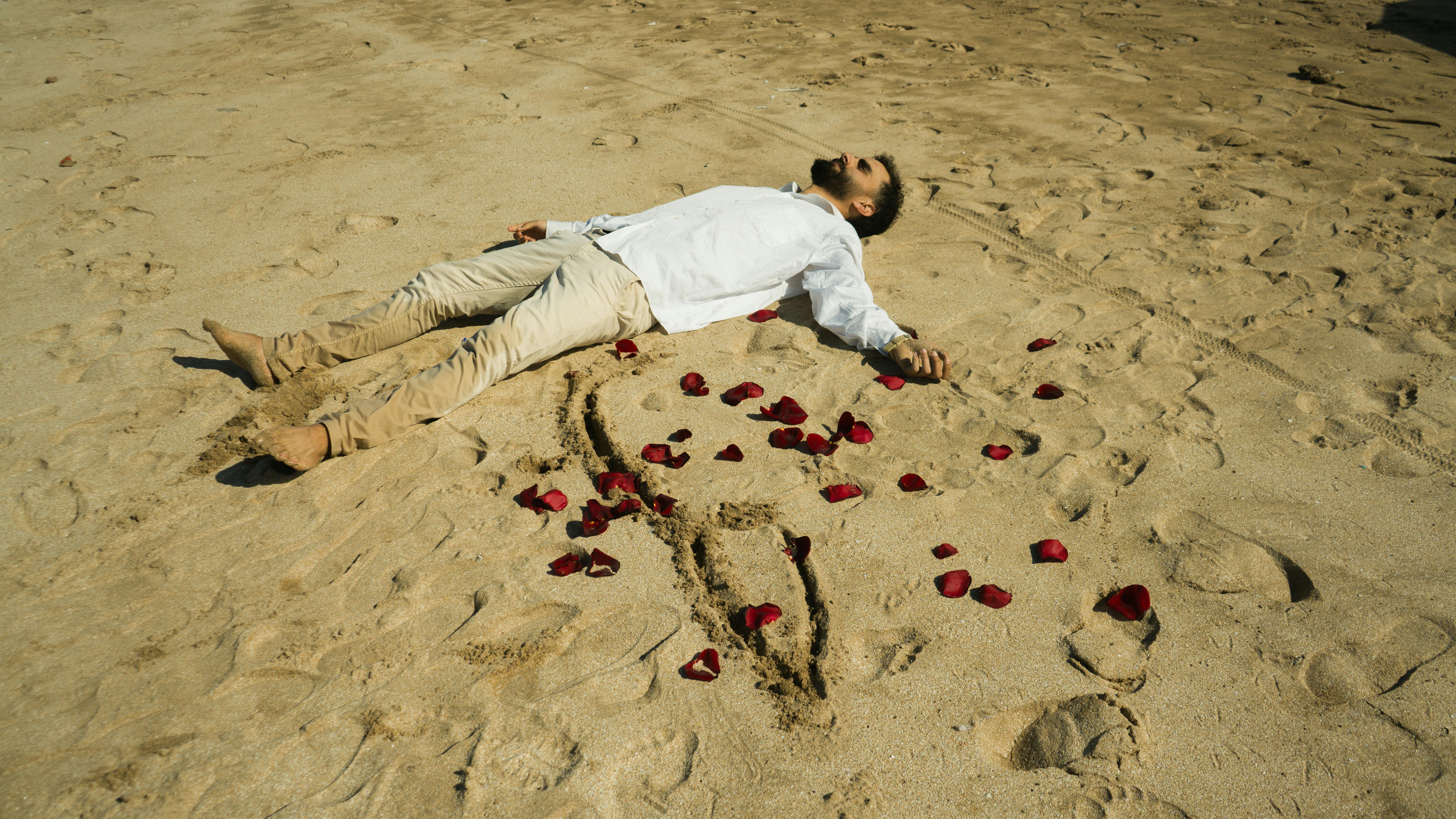 A man in white shirt lies on a sandy beach surrounded by rose petals.