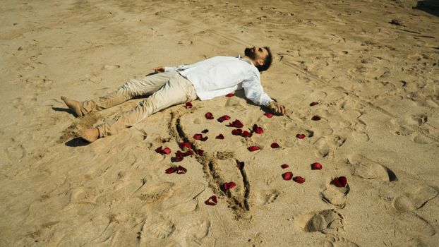 A man in white shirt lies on a sandy beach surrounded by rose petals.