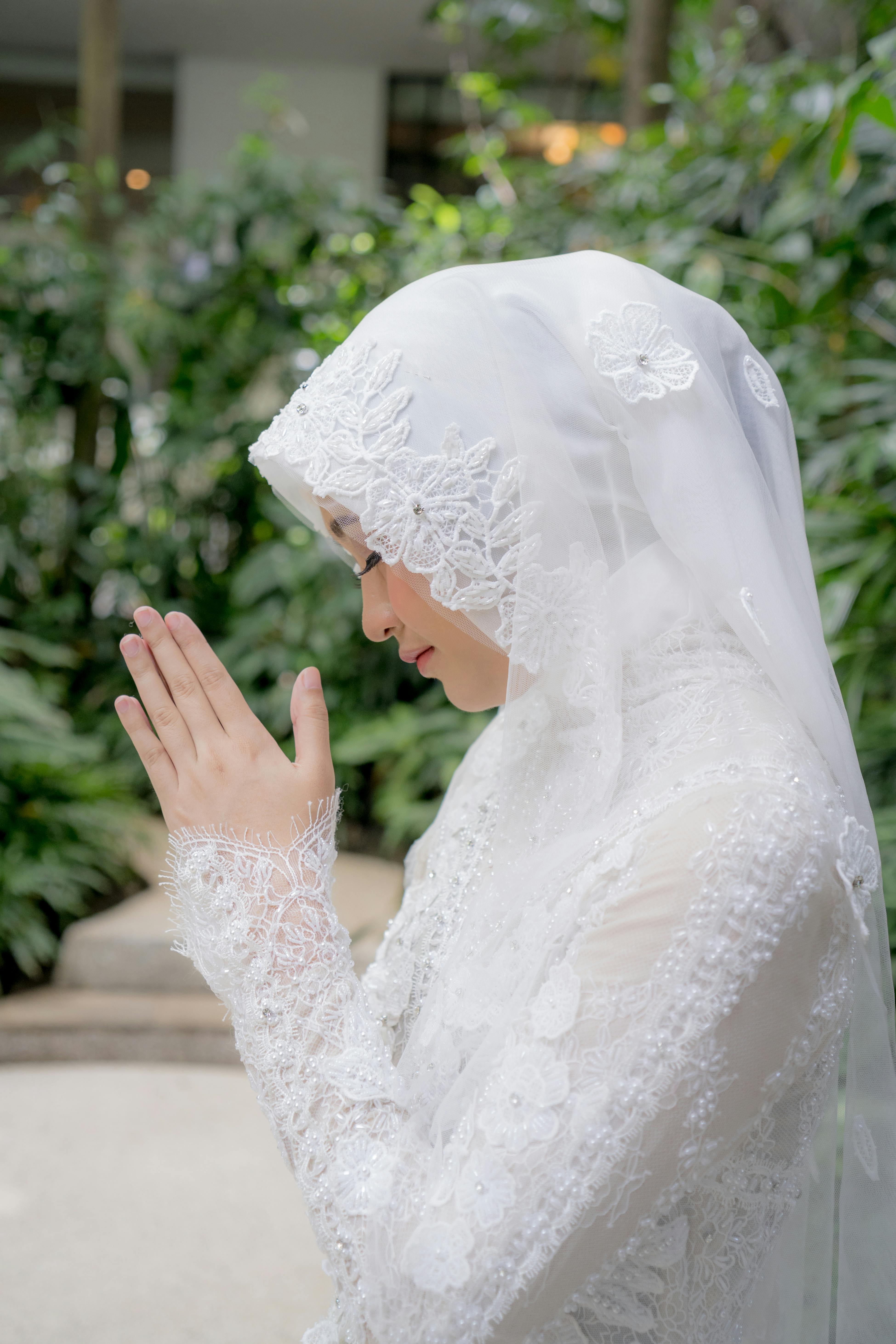 A serene moment of a Muslim woman in a bridal hijab, praying outdoors amidst lush greenery.