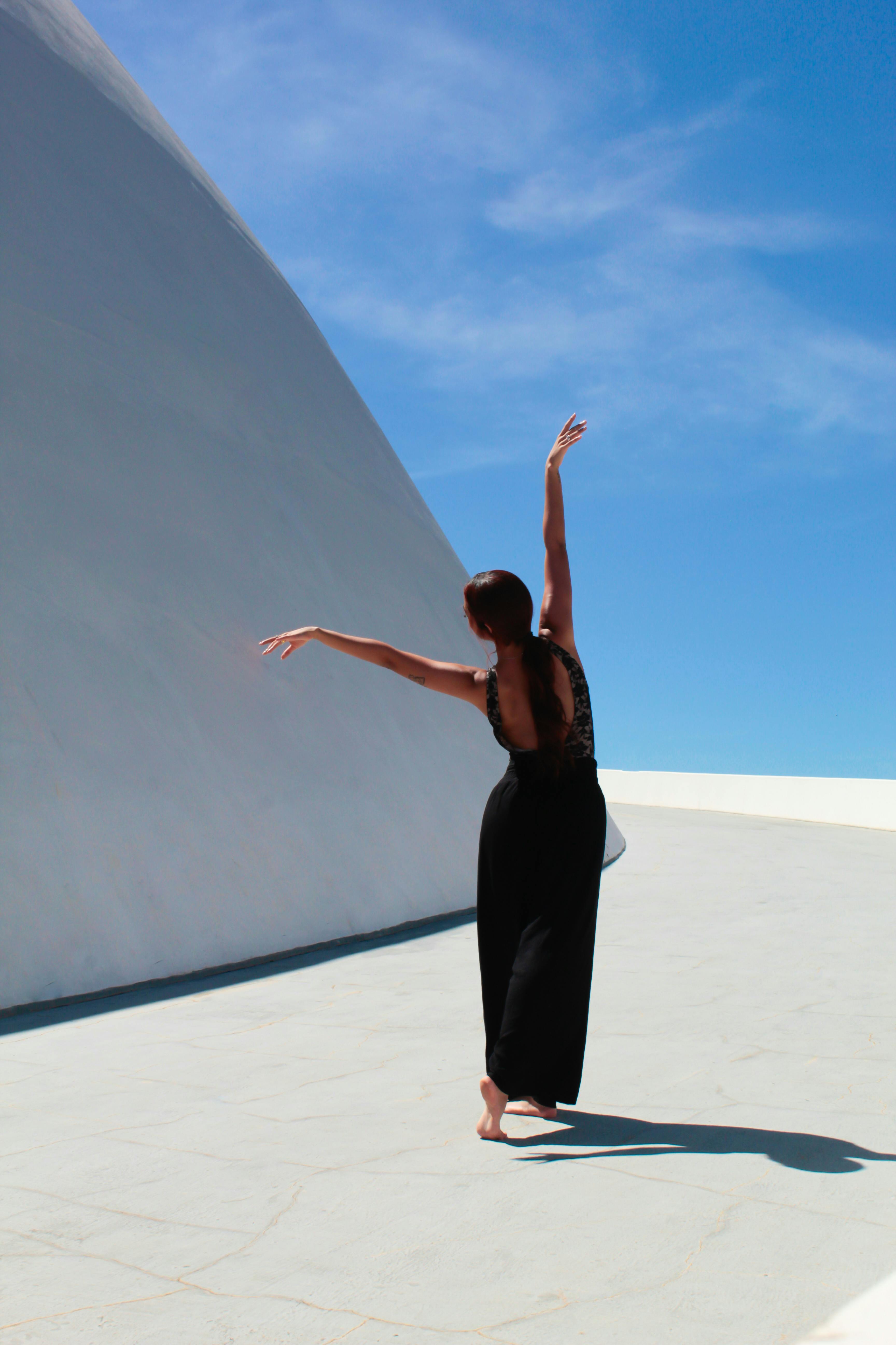 A woman elegantly dances barefoot against a modern architecture backdrop under a clear blue sky.