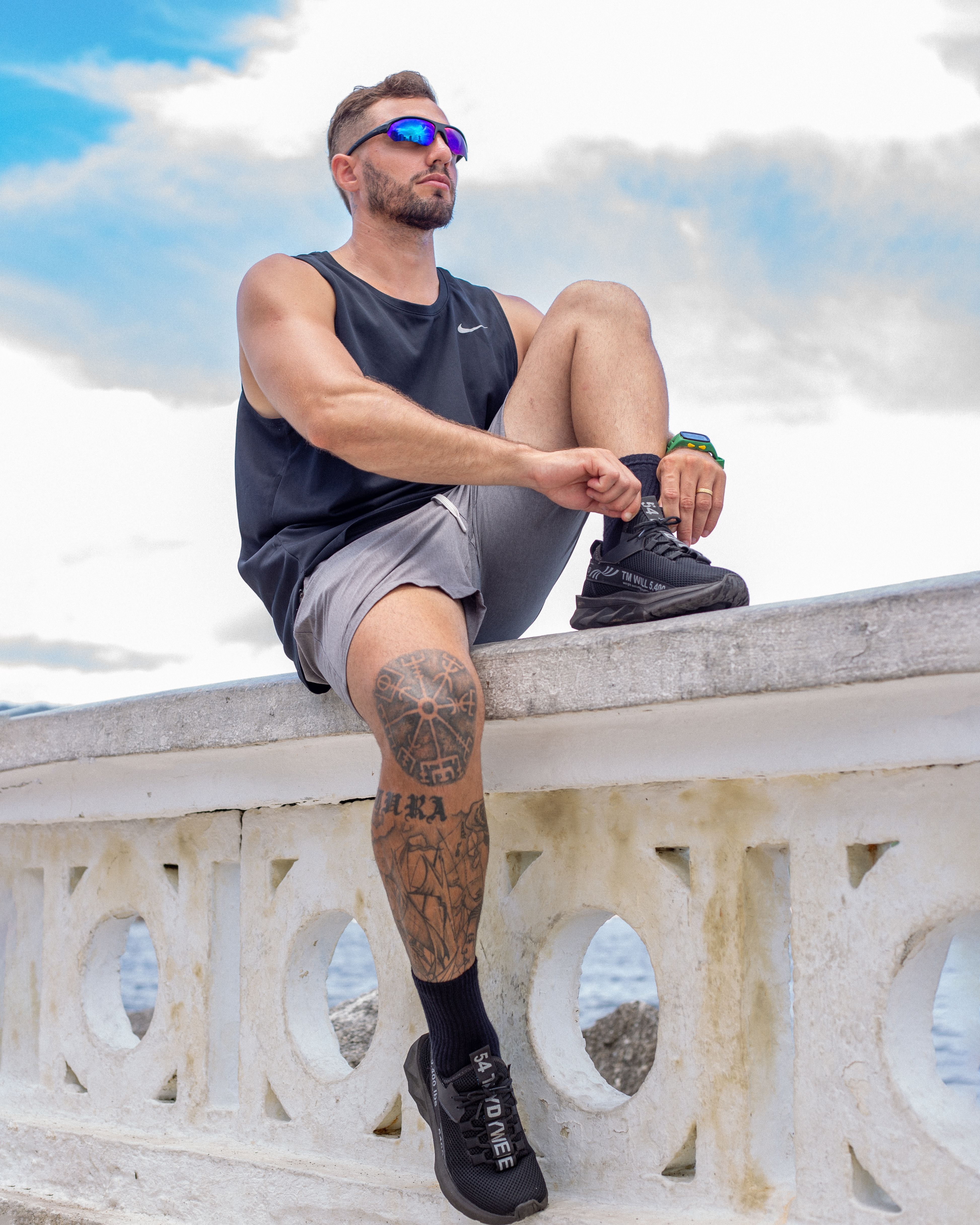 Athletic man with tattoos sits on a ledge outdoors, wearing sporty attire and sunglasses.