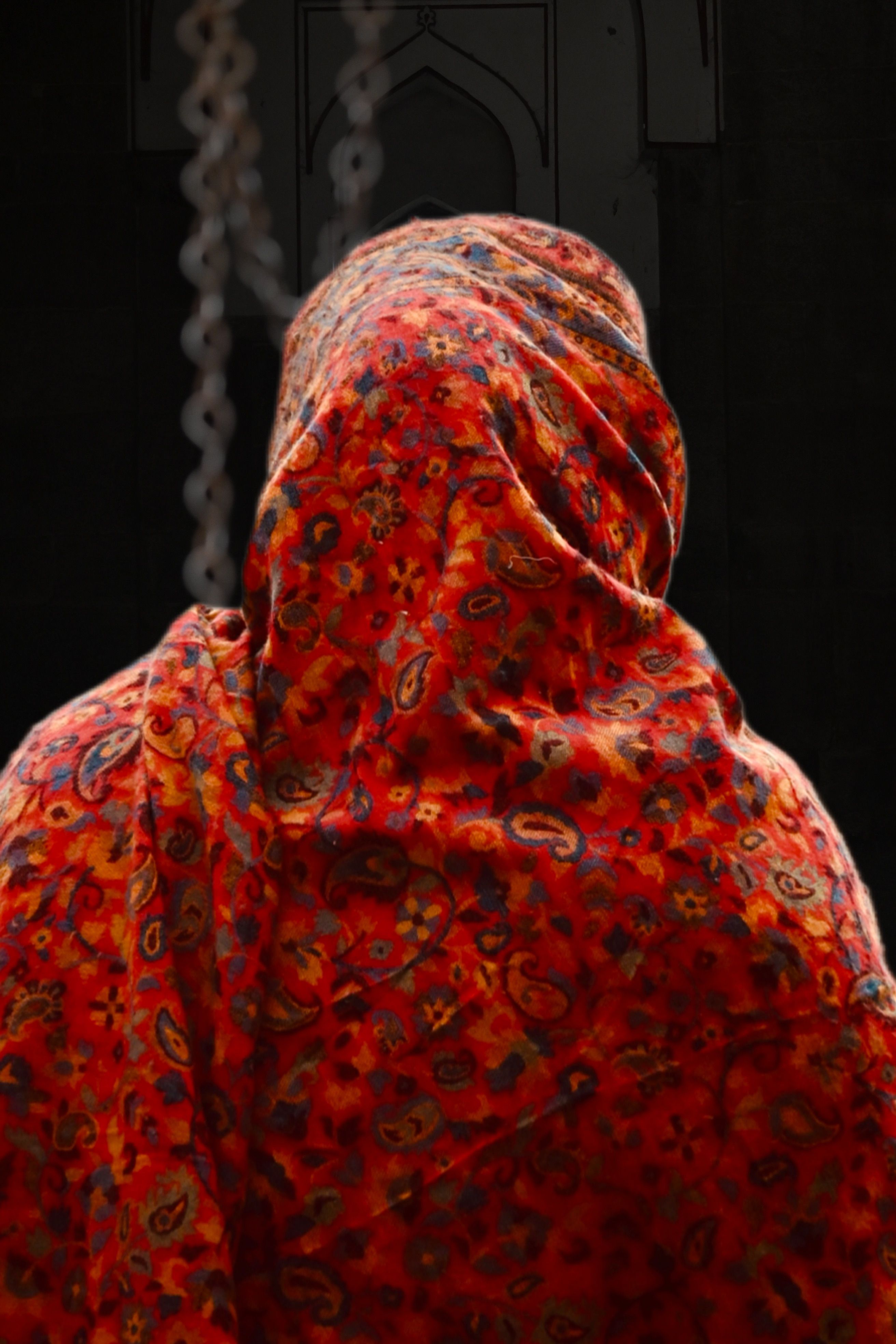 A person draped in a red patterned shawl with intricate designs, captured from behind in an indoor setting.