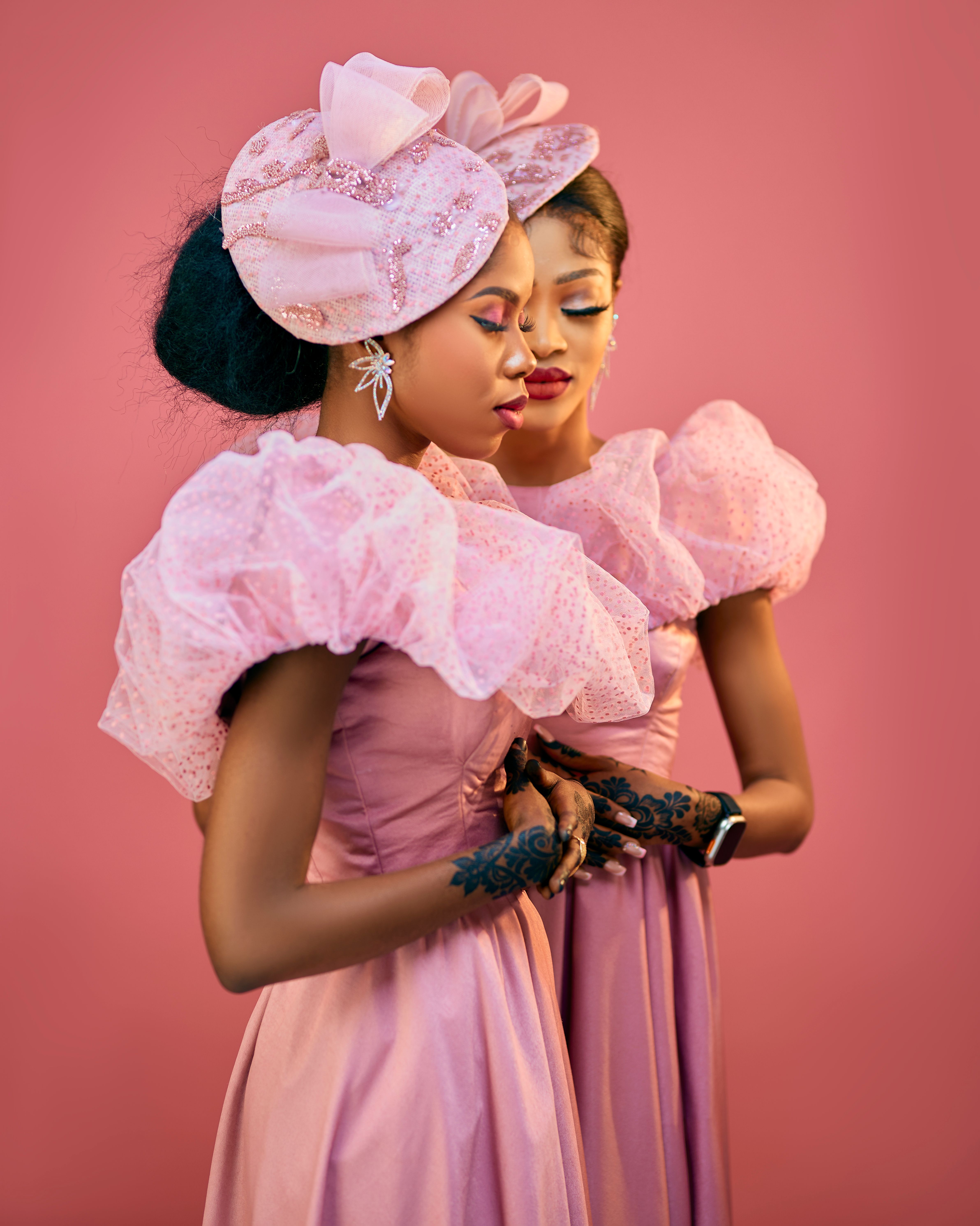 Two women in elegant pink dresses with elaborate headpieces stand gracefully against a pink backdrop.