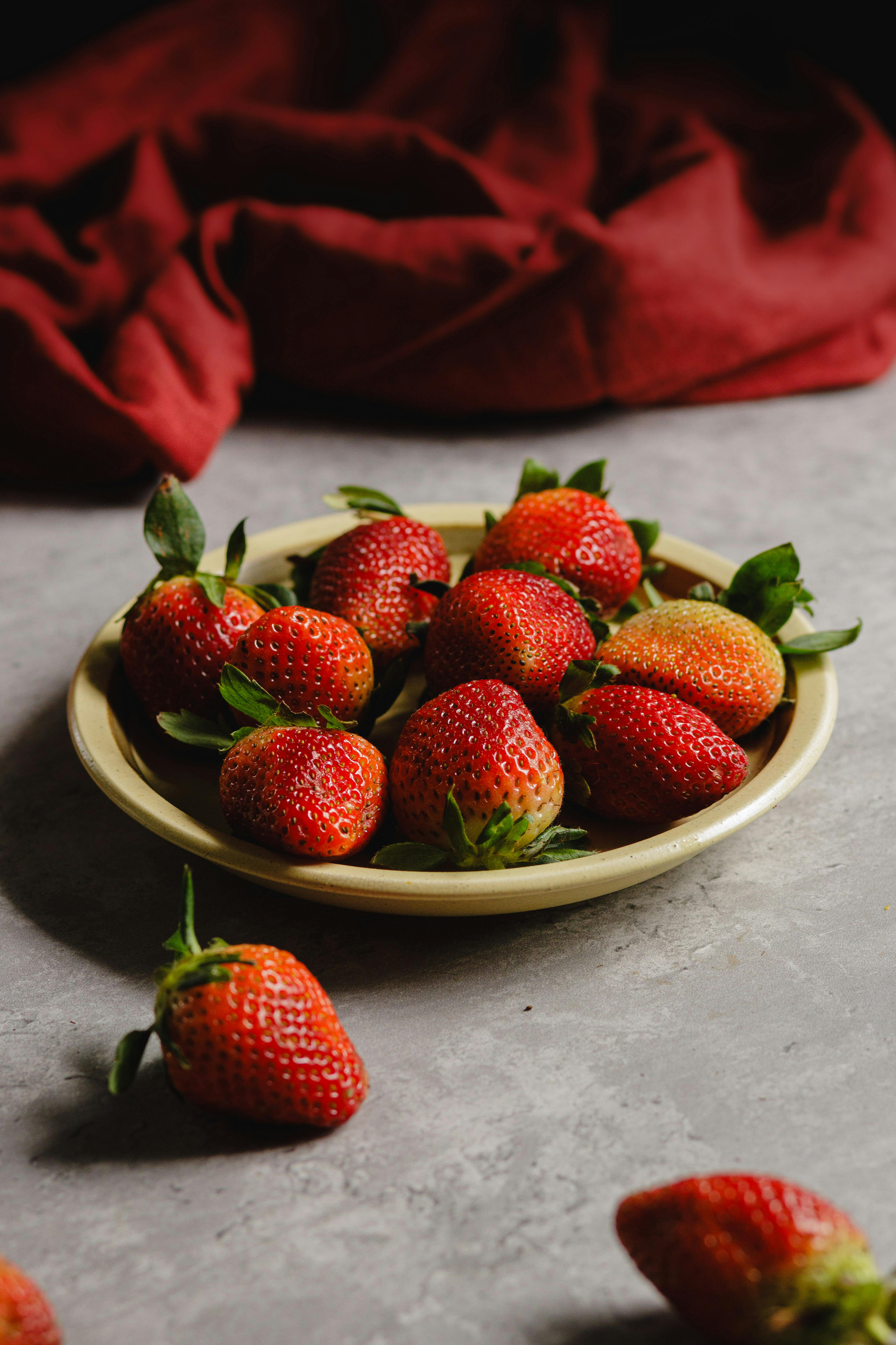 A ceramic plate filled with ripe strawberries on a textured surface, contrasted by a red cloth.