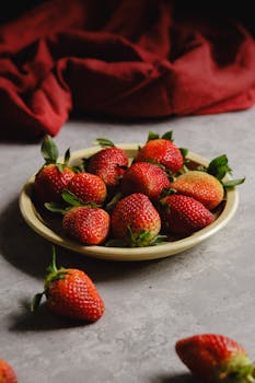 A ceramic plate filled with ripe strawberries on a textured surface, contrasted by a red cloth.