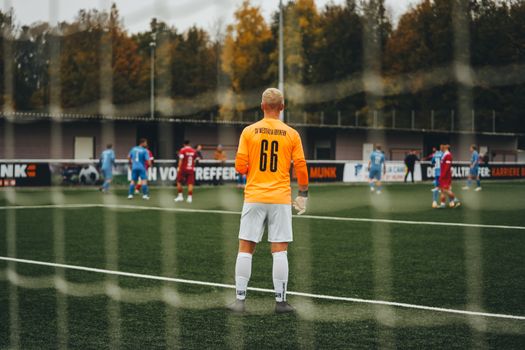 Goalkeeper in orange jersey stands ready during a soccer match on a fall day.