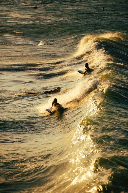 Surfers catch waves during sunset, creating a dramatic seascape with golden hues.