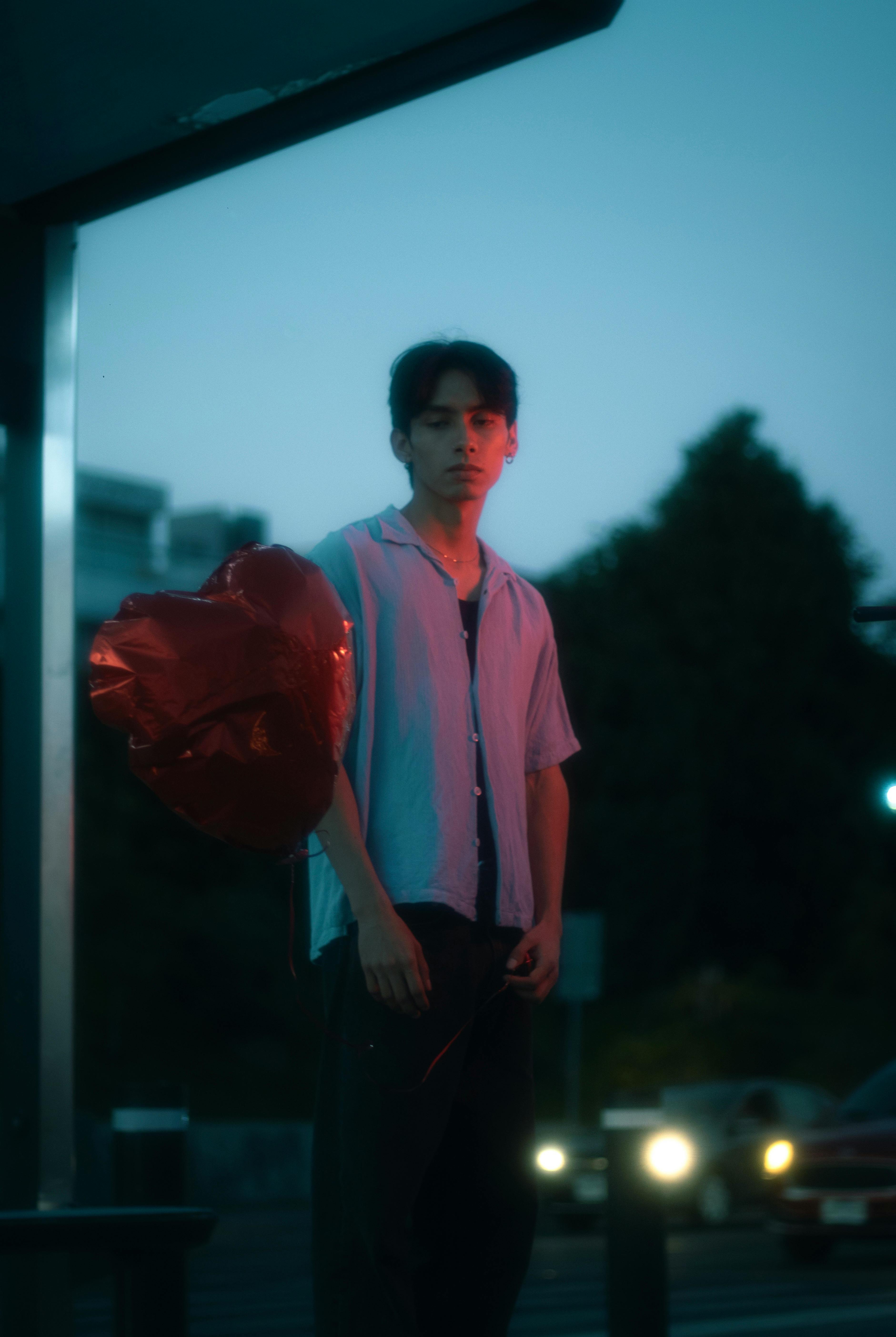 A young man stands outdoors at night holding a heart-shaped balloon, evoking emotions.