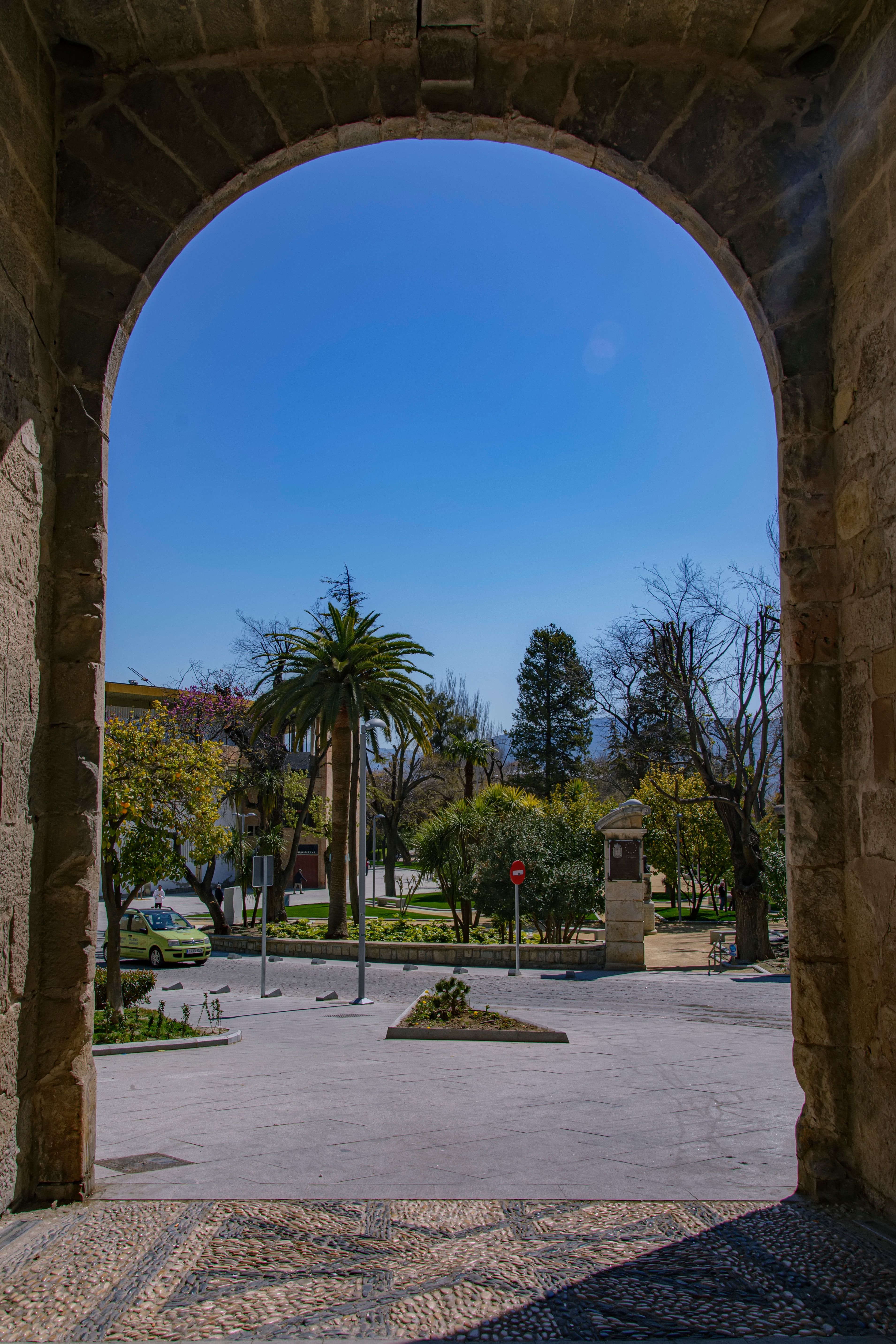 Fotomural Explora Jaén a través de un arco histórico, capturando una escena tranquila del parque bajo un cielo azul claro.