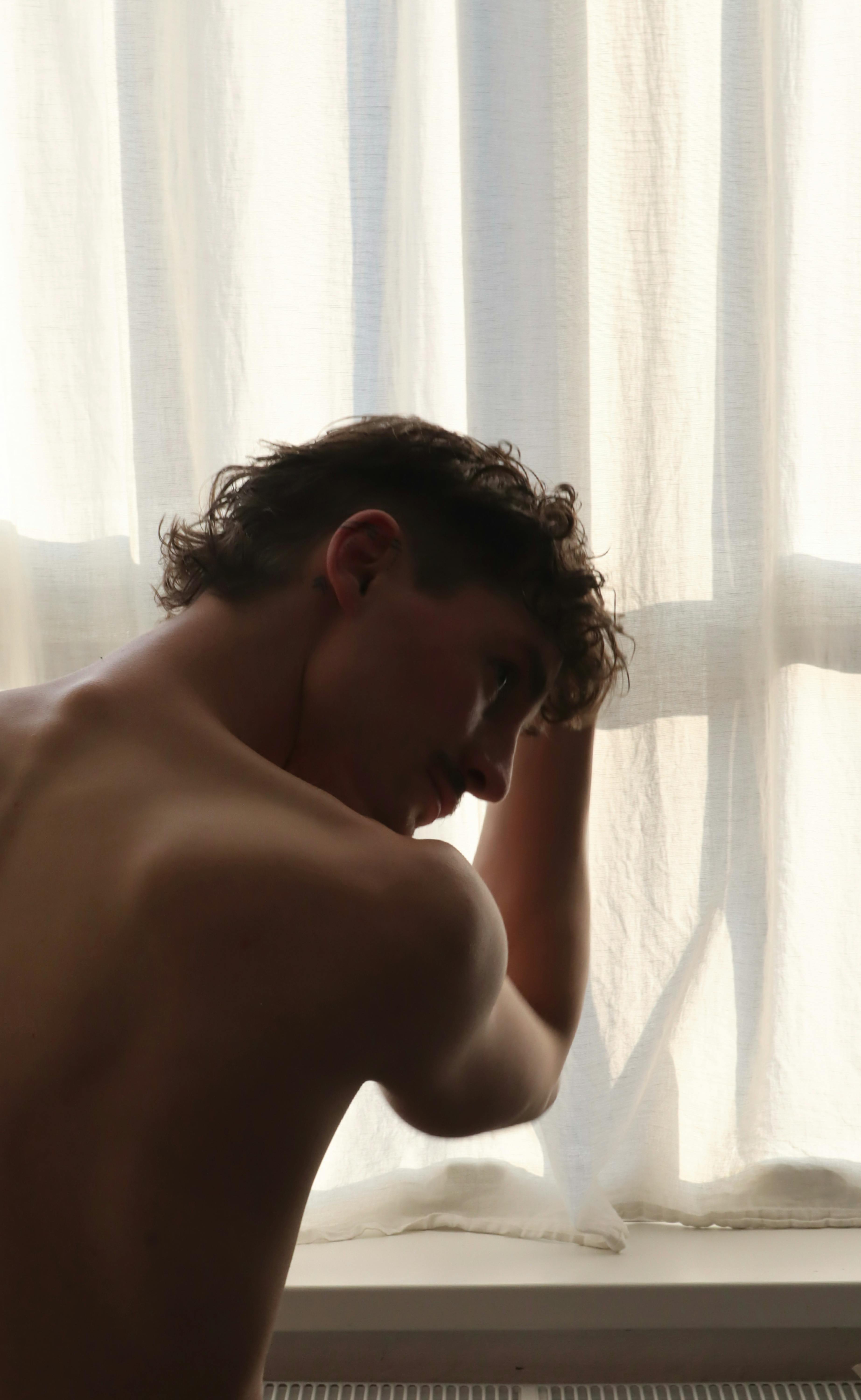 bezplatná Siluetovaný muž bez košile vypadá z okna, měkké osvětlení evokuje introspekci. Základová fotografie