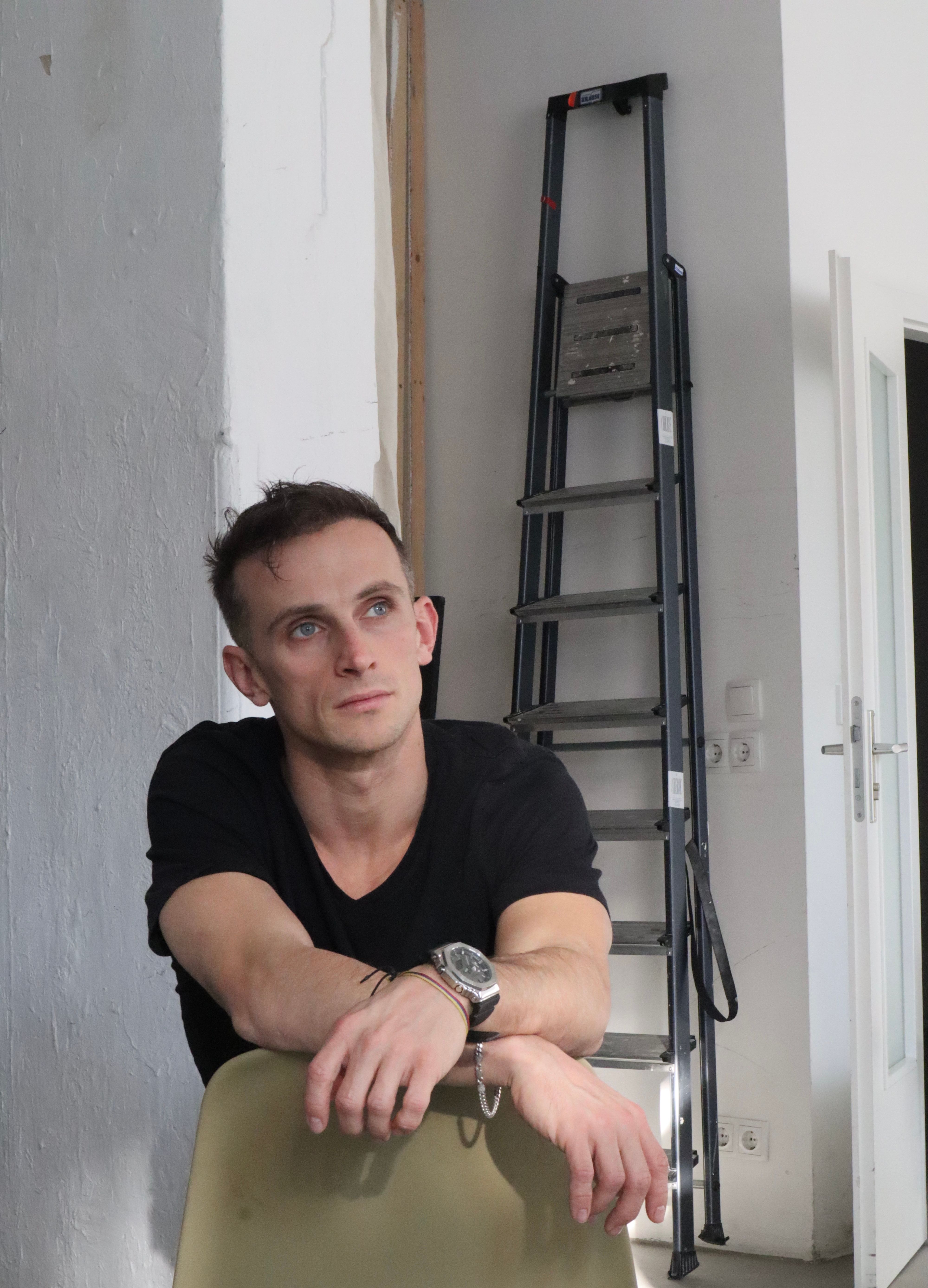A young man sits indoors looking thoughtful, next to a ladder.