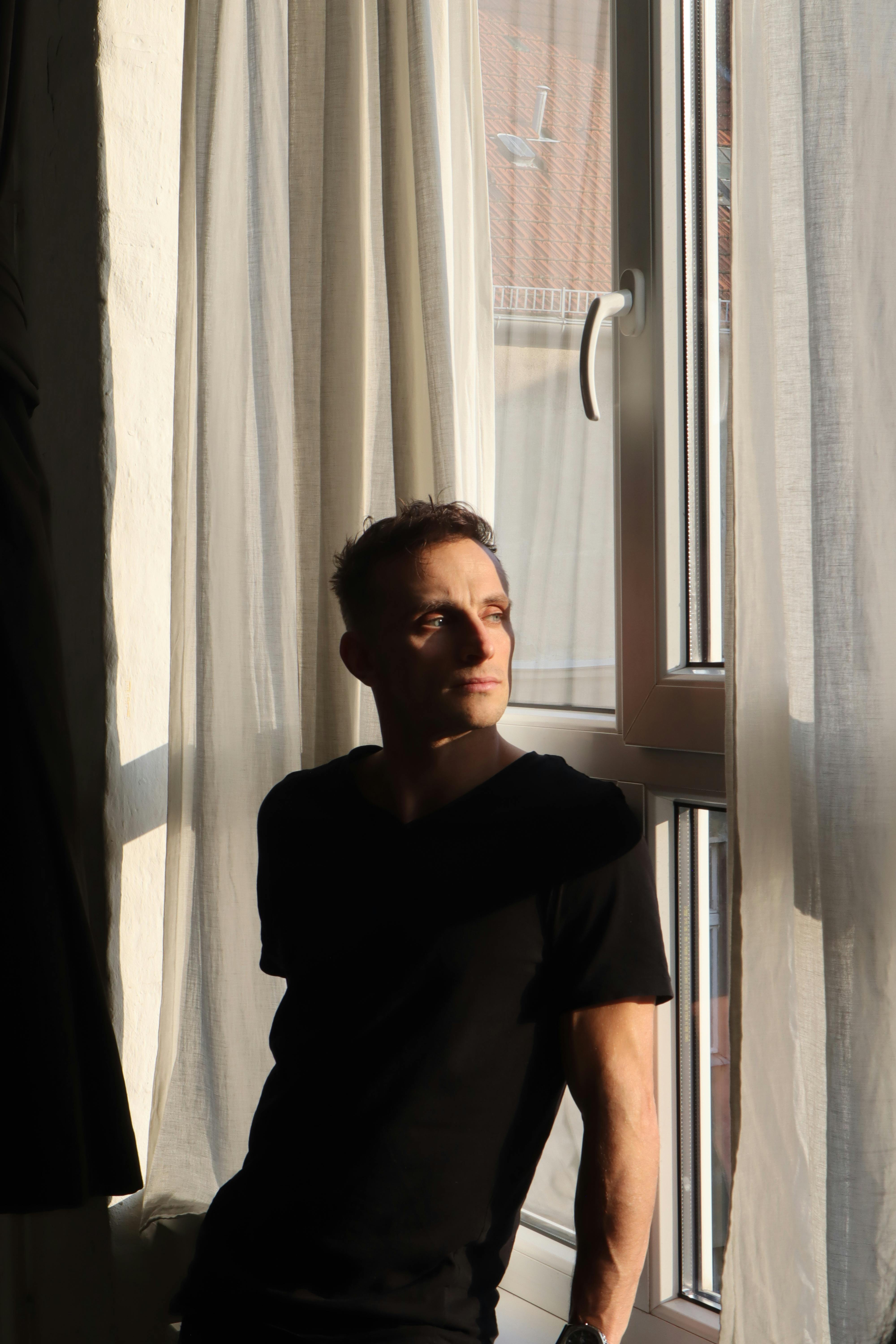A young man gazes thoughtfully by a window in Berlin, surrounded by soft light and shadows.