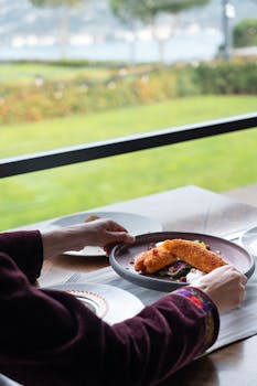 Person enjoying a plated meal by a window with a lake view.
