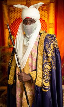 Portrait of a man in traditional African tribal attire holding a spear indoors.