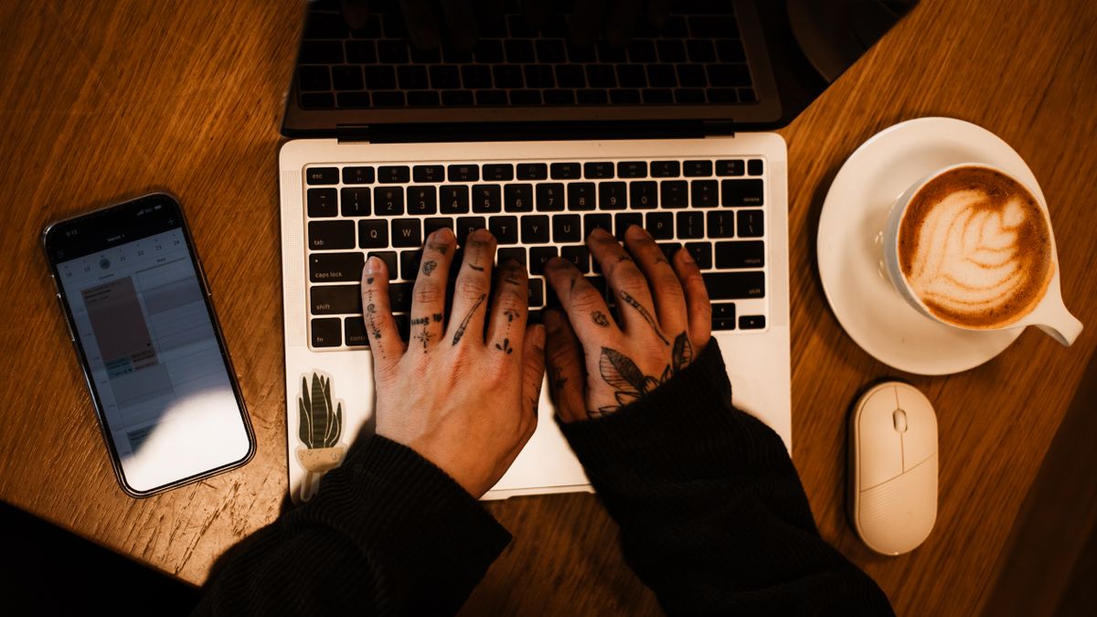 タトゥーの入った手でノートPCを操作している、コーヒーのある机のイメージ