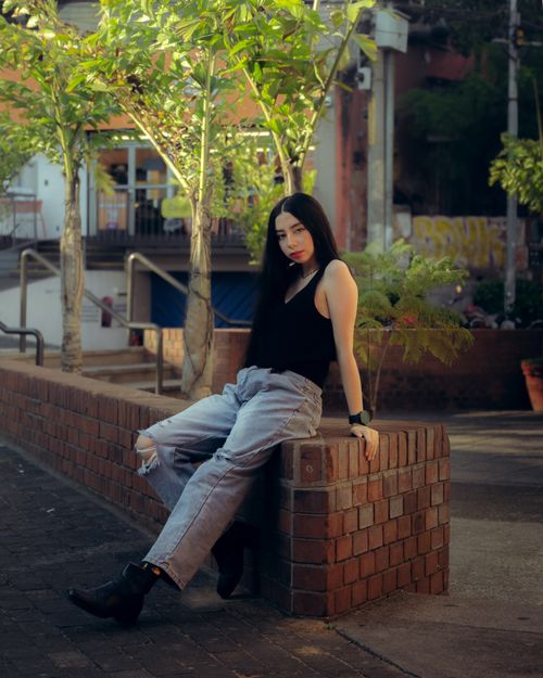 Free A stylish woman poses outdoors on a brick wall surrounded by greenery and urban elements. Stock Photo