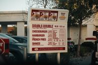 In-N-Out drive-thru menu featuring burgers, fries, and shakes under a California sky.
