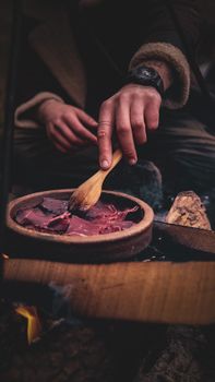A person cooking meat outdoors using a wooden spatula over an open fire.