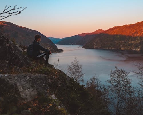 Man Sitting on Cliff Facing Calm Body of Water