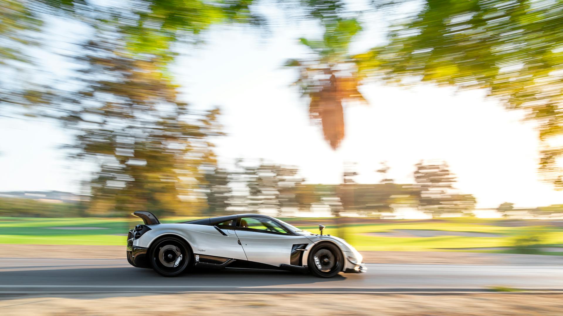 Silver sports car speeding along a road with dynamic motion blur and sunlit trees.