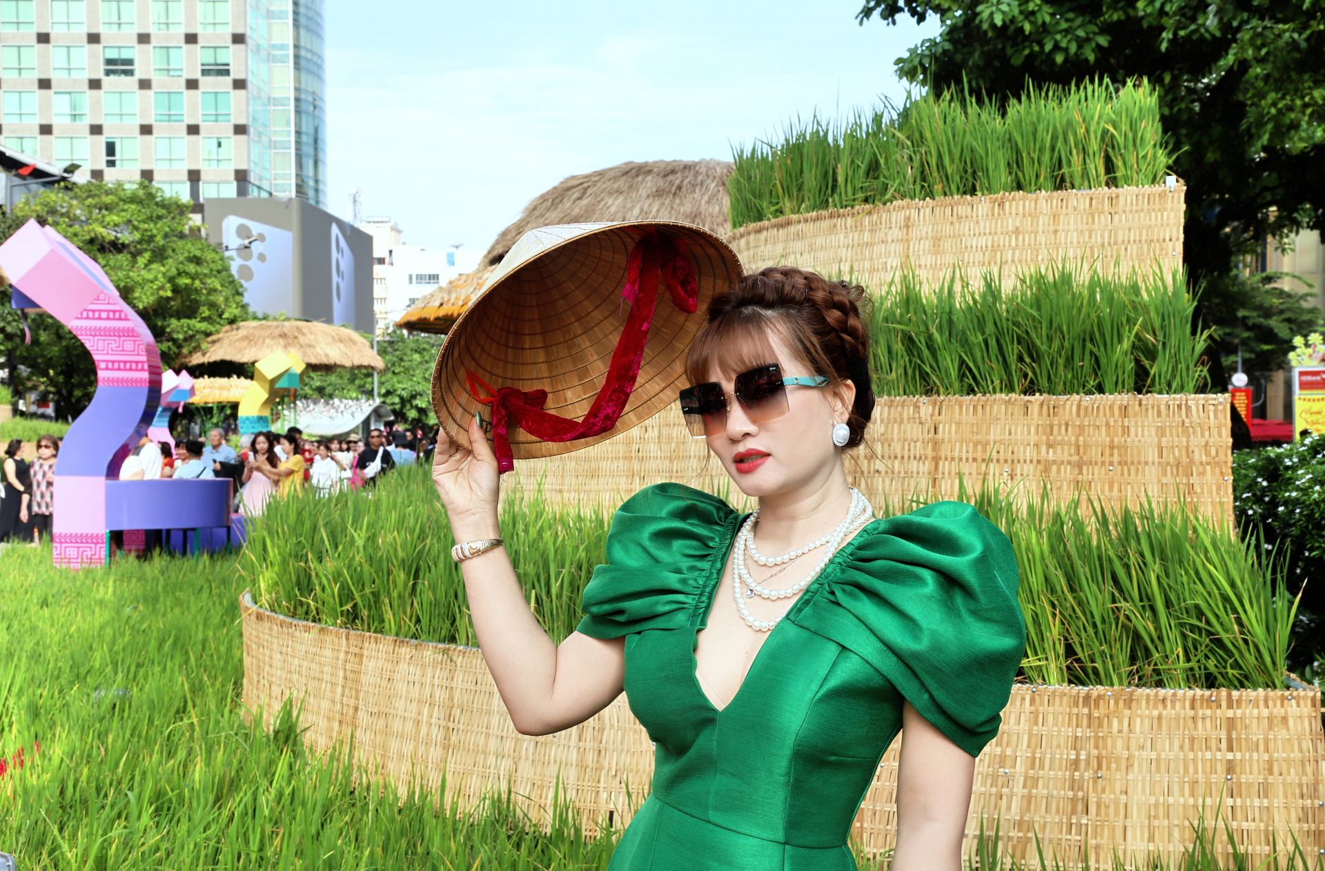 Stylish woman in green dress holding conical hat in a vibrant, cultural outdoor setting.