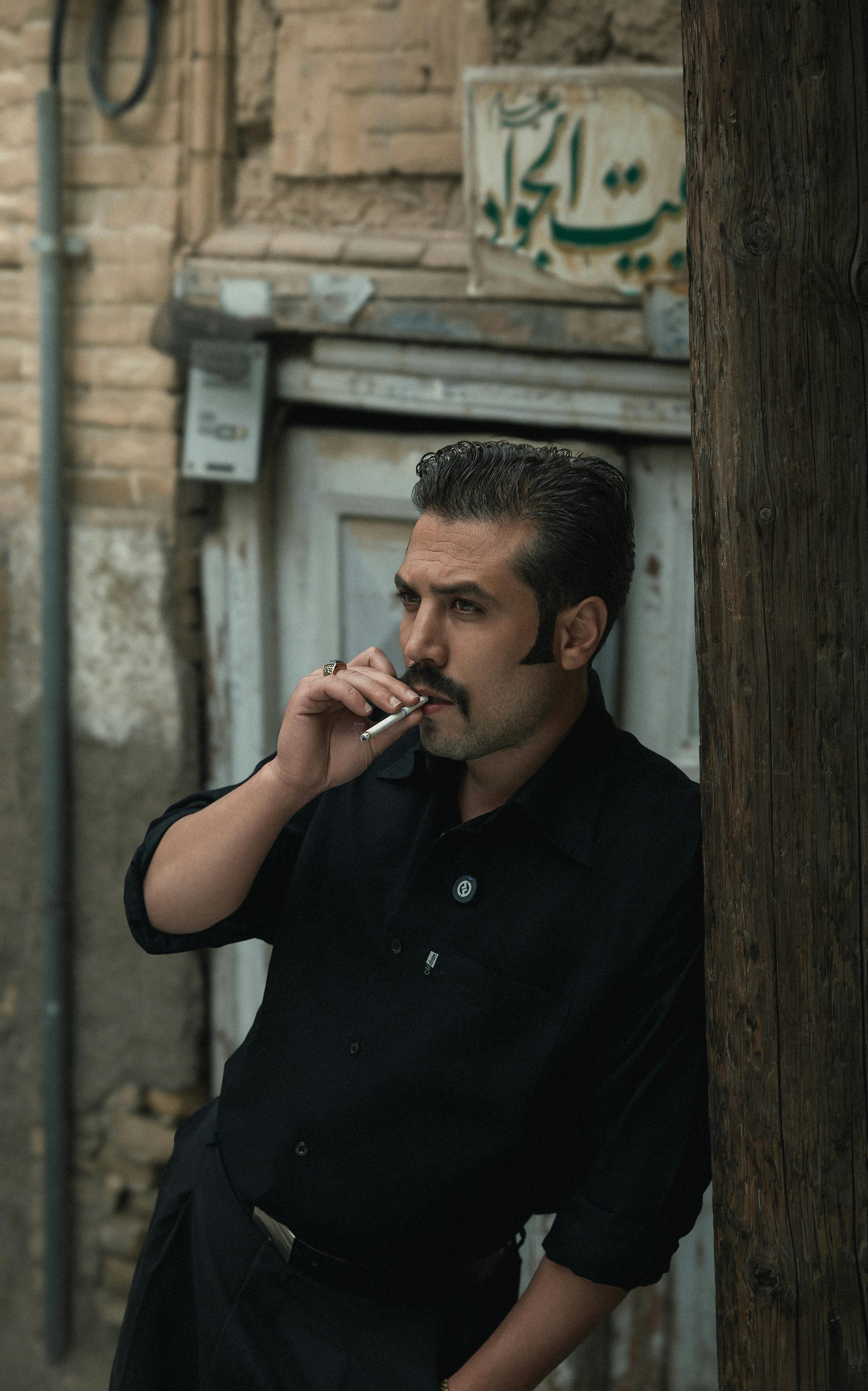 Man leaning on a post in urban alley, smoking a cigarette.