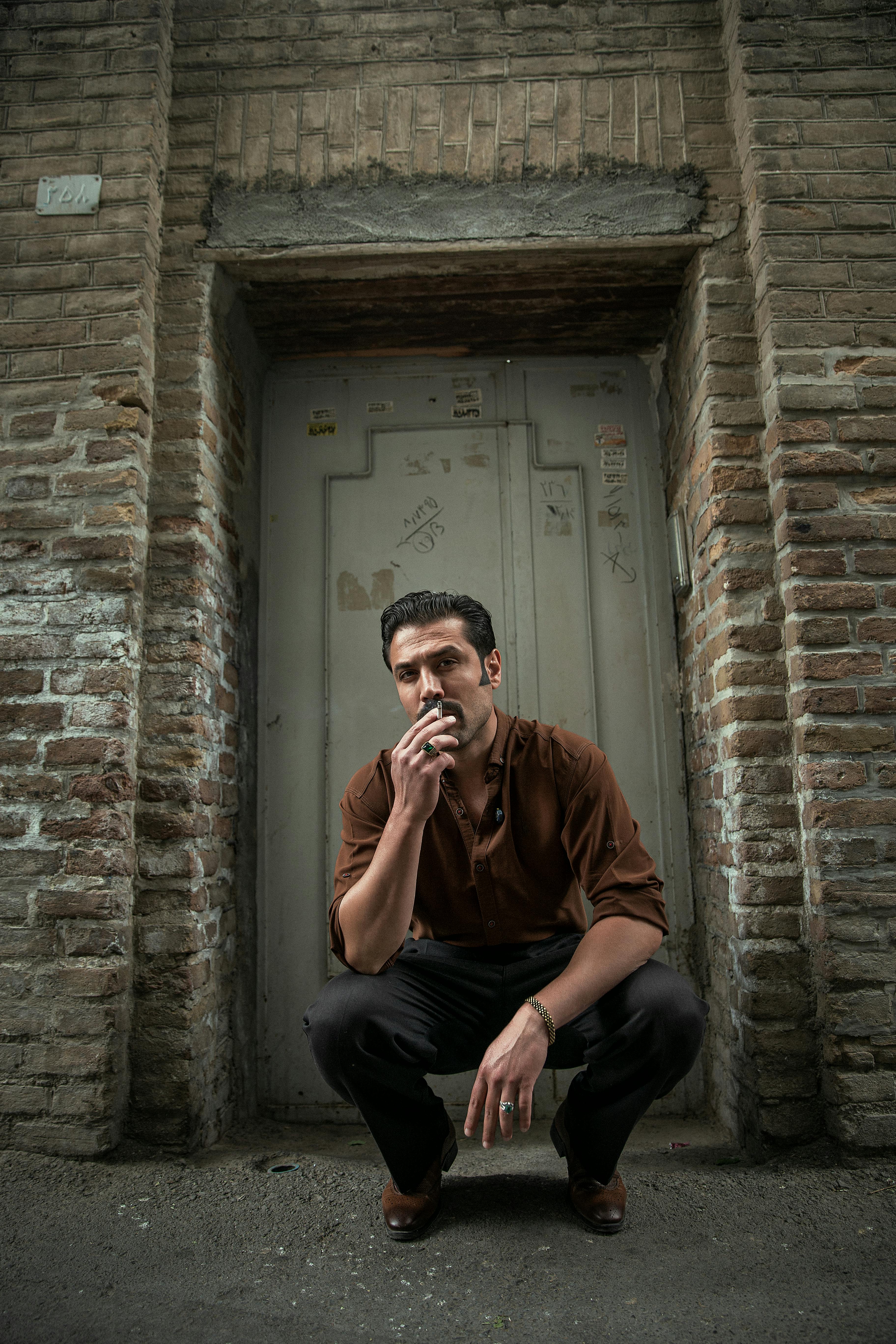 Man squatting against a rustic brick wall, thoughtful and casual in urban setting.