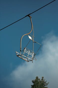 A solitary chairlift suspended against a blue sky with clouds and treetops.