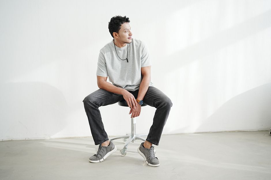 A man in casual attire sitting on a stool in a well-lit room.