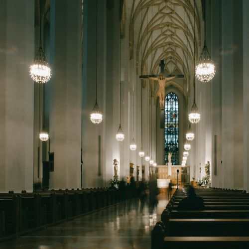 Free Gothic church interior showcasing ornate chandeliers and stained glass window with warm, inviting ambiance. Stock Photo