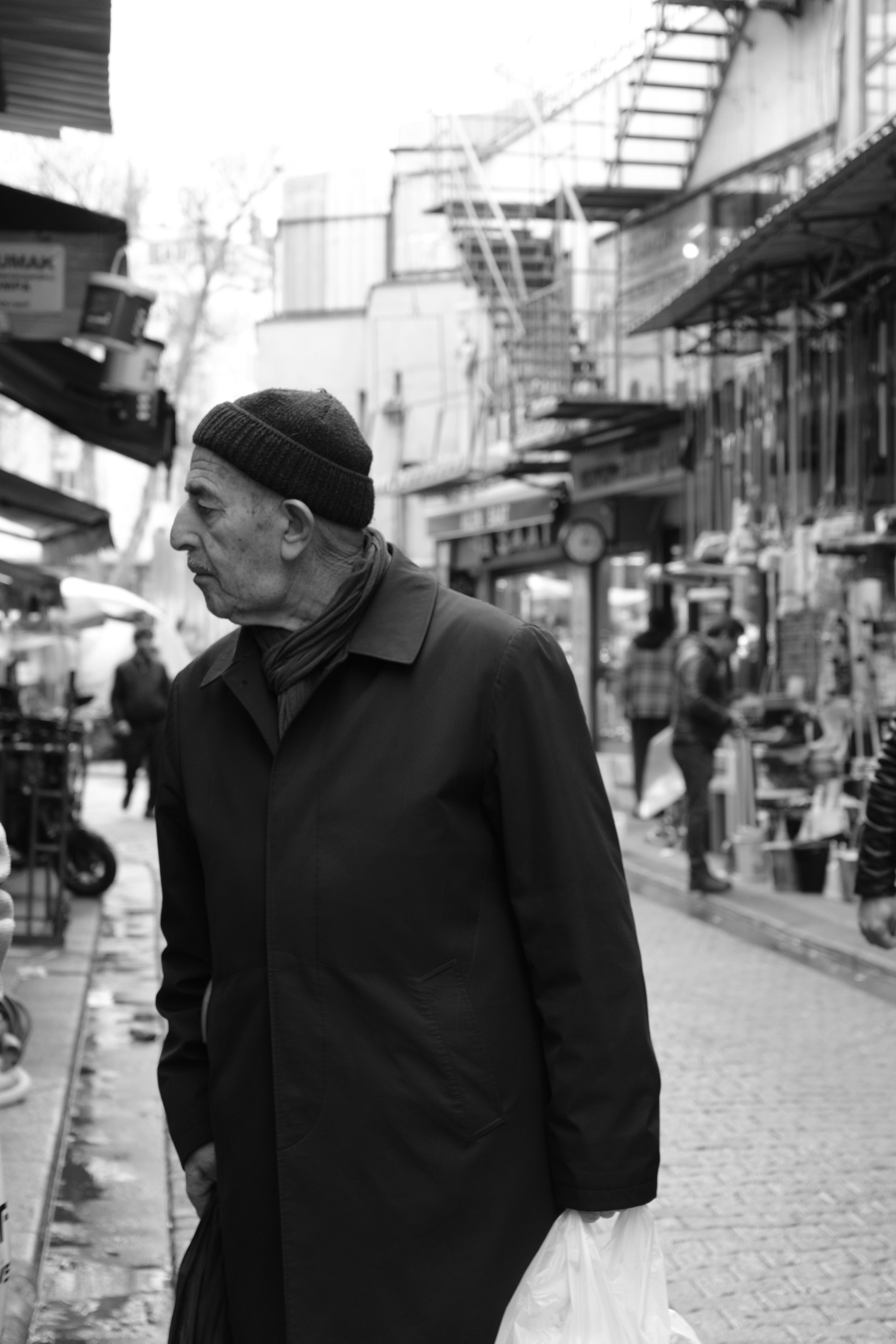 A black and white photo of an elderly man in a beanie walking through a vibrant street market.
