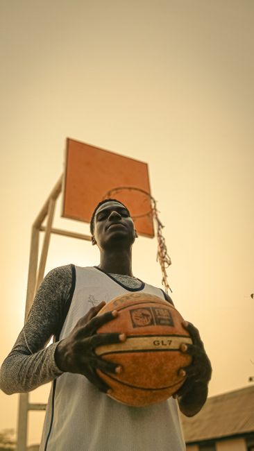 Teen basketball player holding ball under outdoor hoop during day.