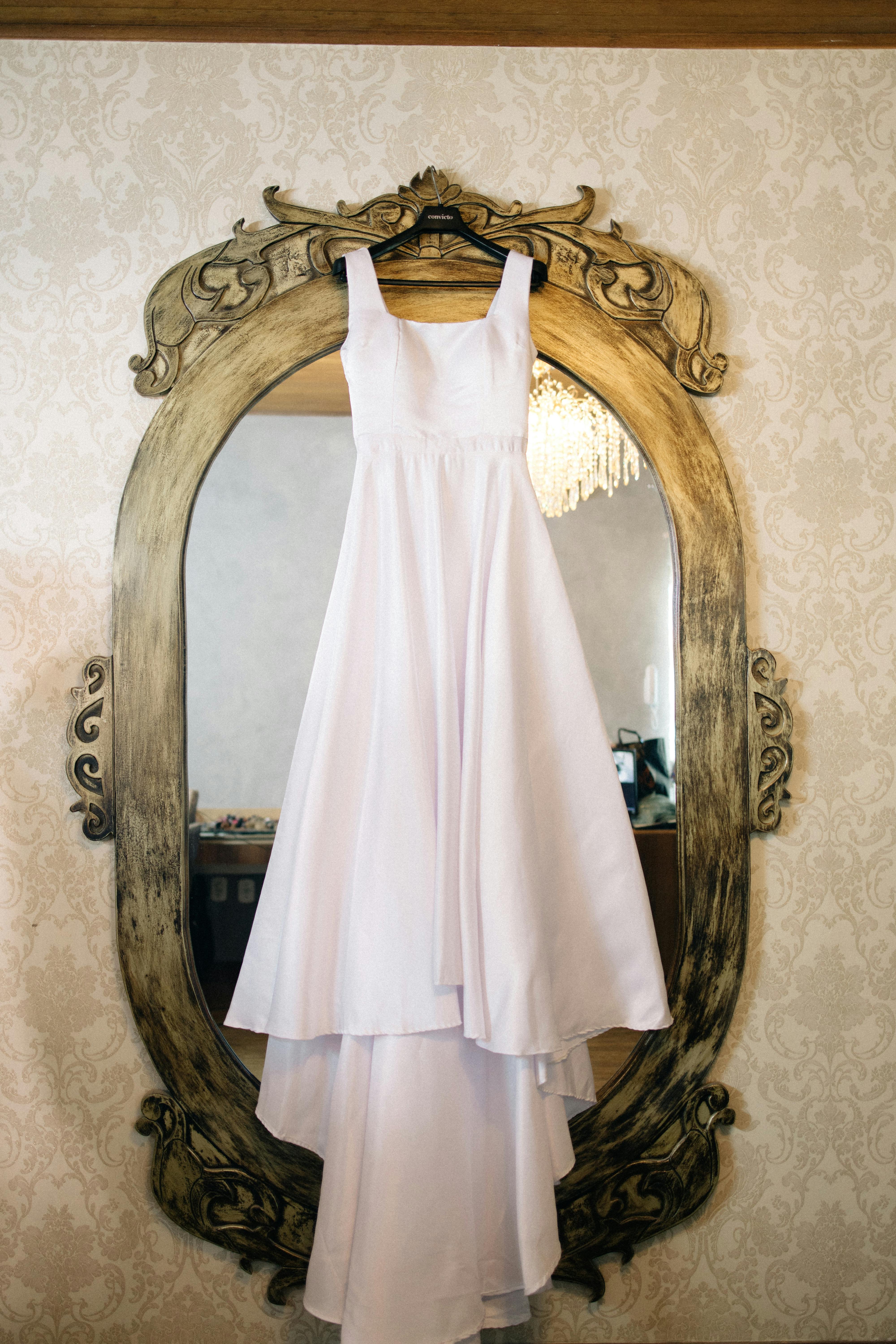 Gratis Un vestido de novia blanco cuelga elegantemente de un espejo vintage adornado en una habitación bien iluminada. Foto de stock