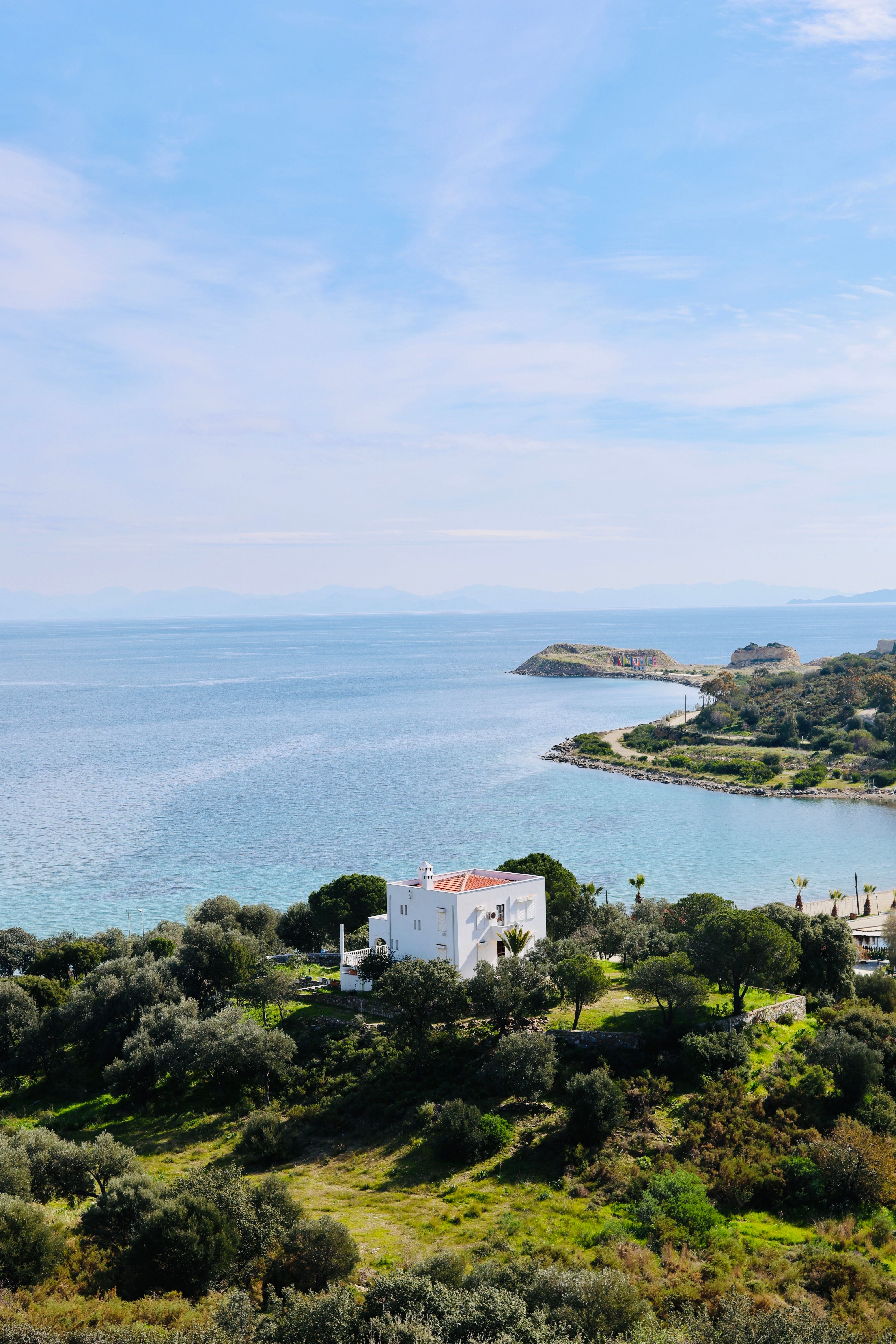 A serene view of the coastline in Muğla, Türkiye, featuring lush greenery and a white villa under a clear blue sky.
