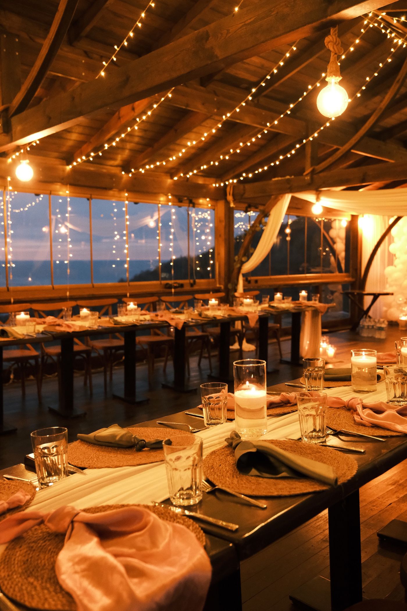 A long dinner table laid with candles under a timber roof hung with lights