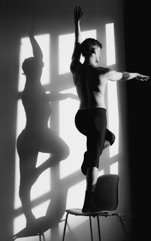 Artistic black and white photo of a dancer balancing on a chair with shadow play on the wall.