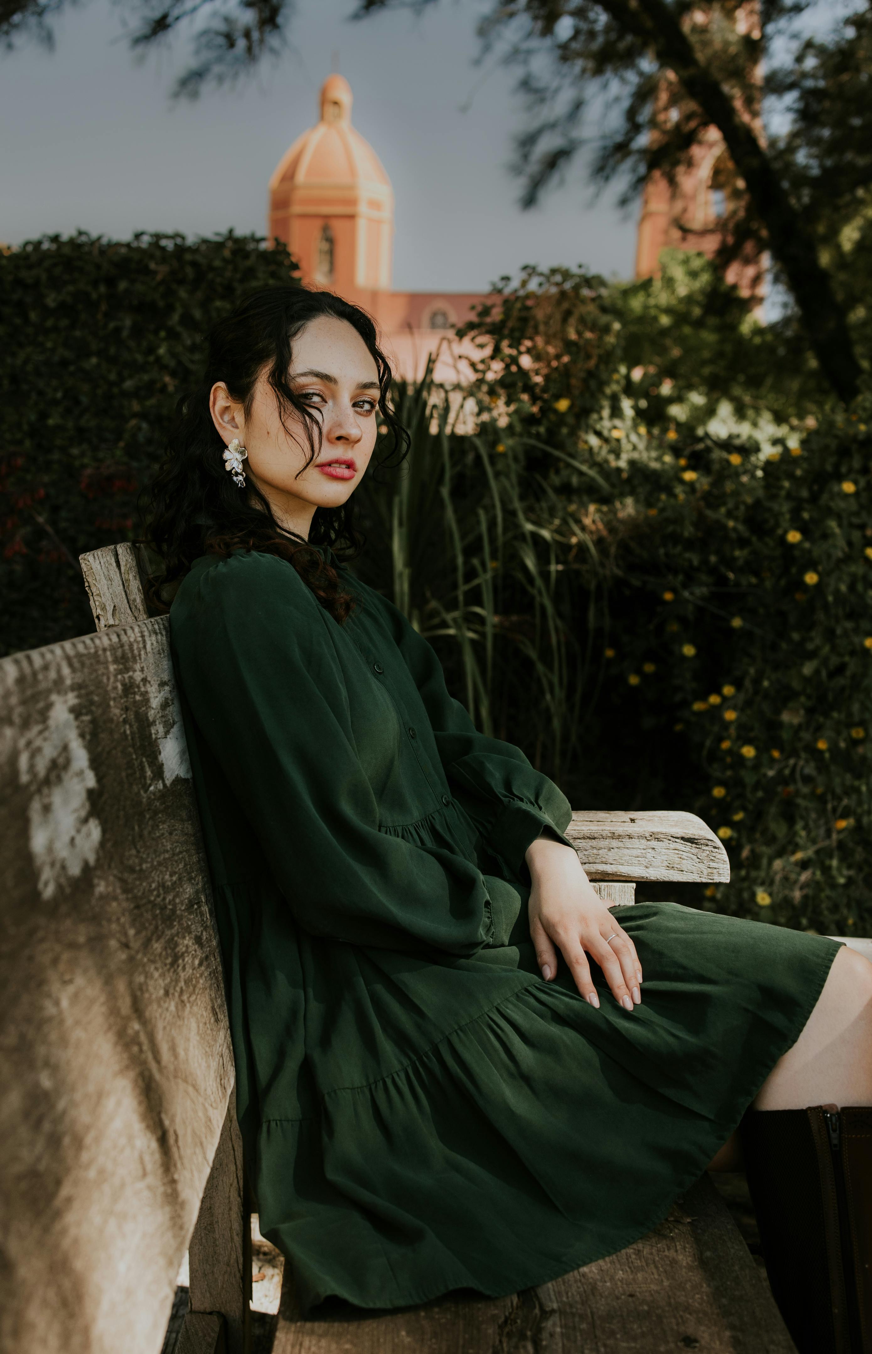 A woman in a green dress sits on a bench near a historical building outdoors.