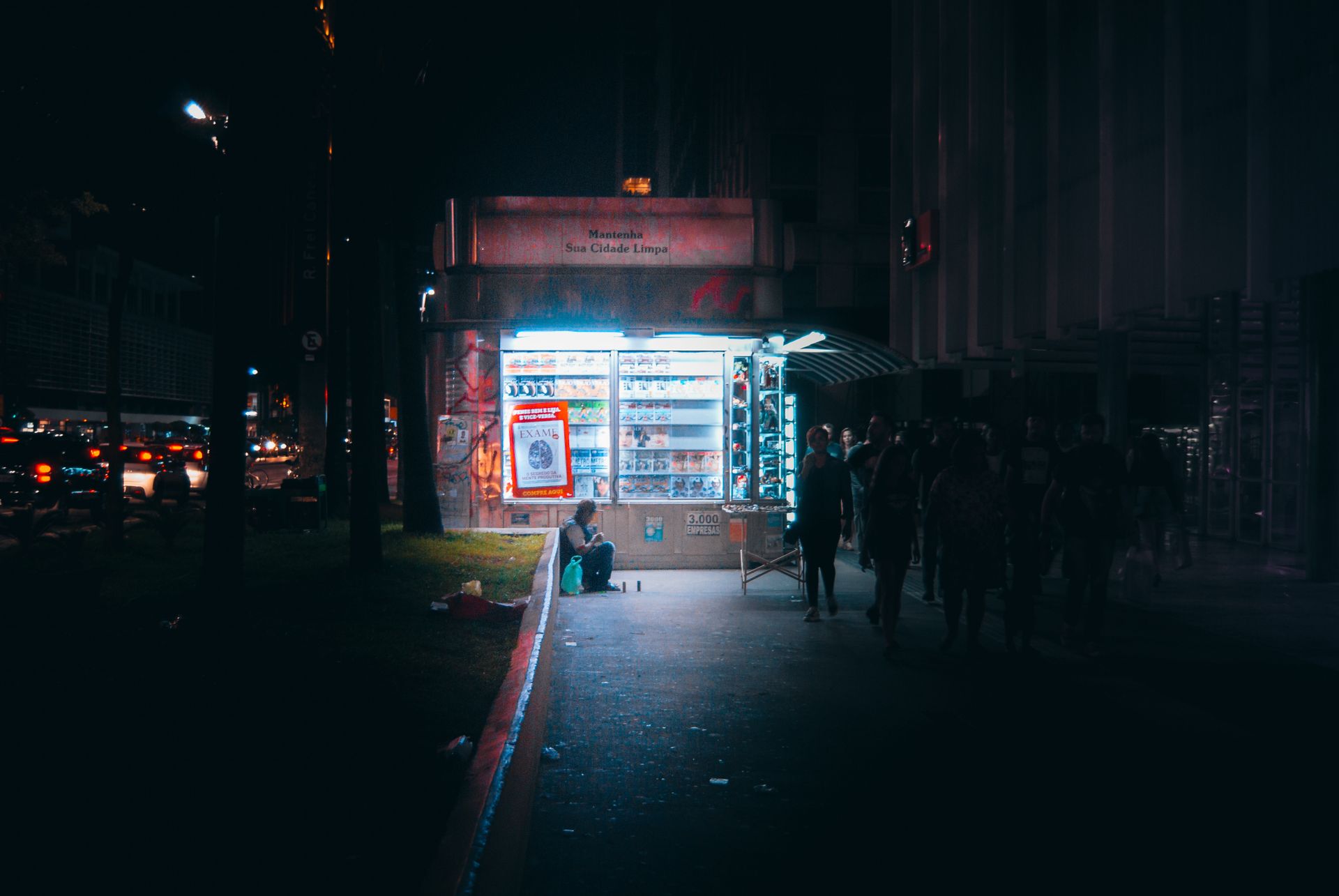 A nighttime street scene featuring an illuminated kiosk surrounded by people and city lights.