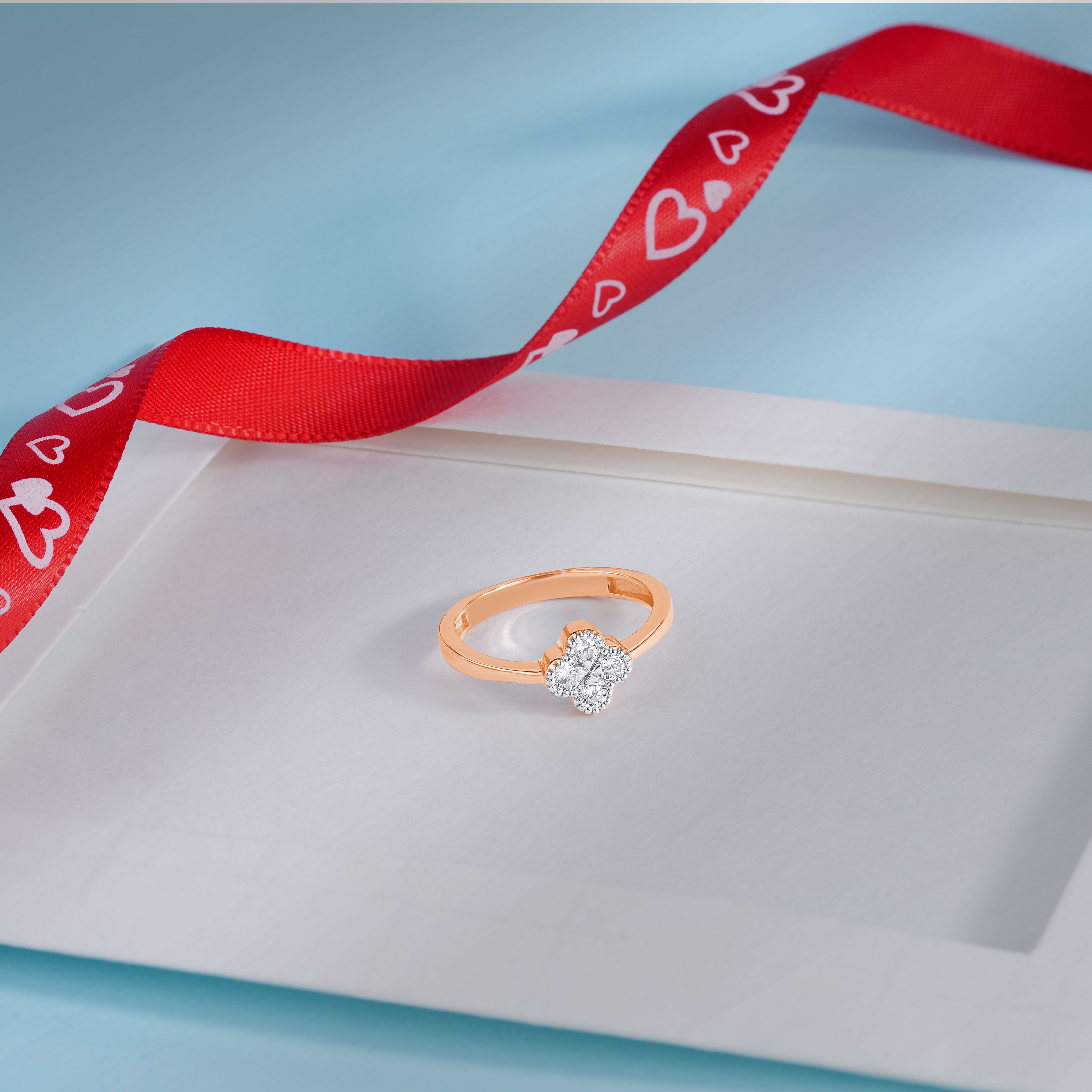 A beautiful diamond ring placed on a white surface with red heart-patterned ribbon in elegant layout.